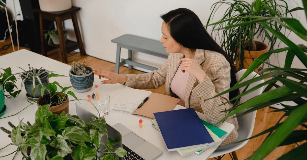 Une femme en manteau marron travaille à un bureau rempli de plantes avec un ordinateur portable et des cahiers.