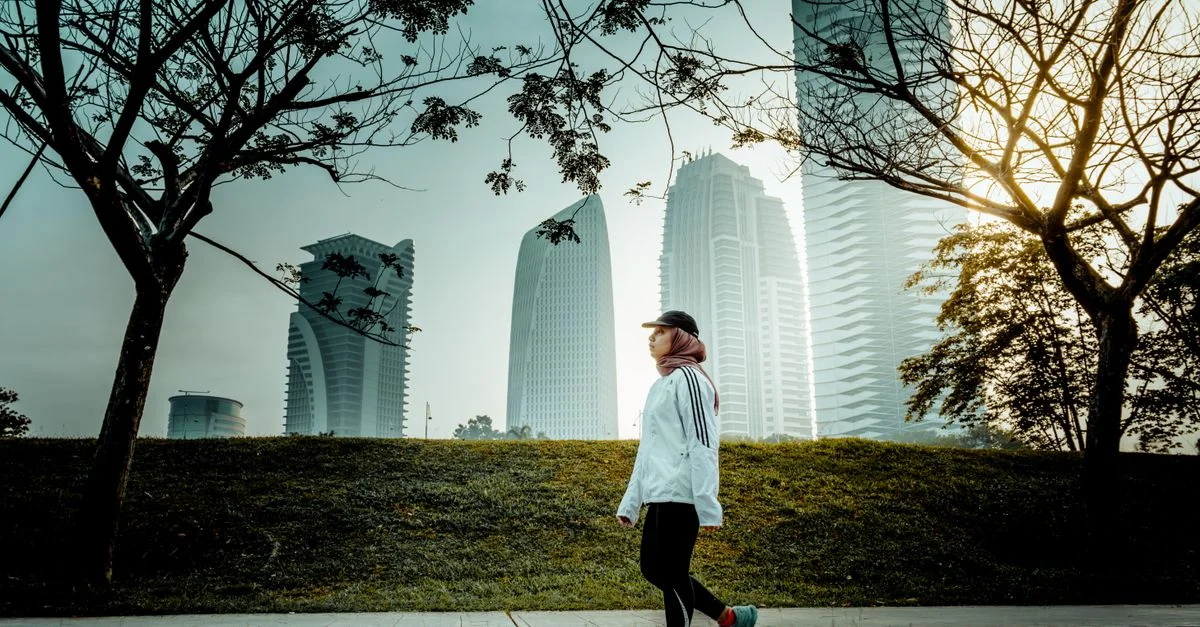 Une femme se promène dans un parc de Putrajaya, entourée d'arbres avec des gratte-ciel urbains en arrière-plan.