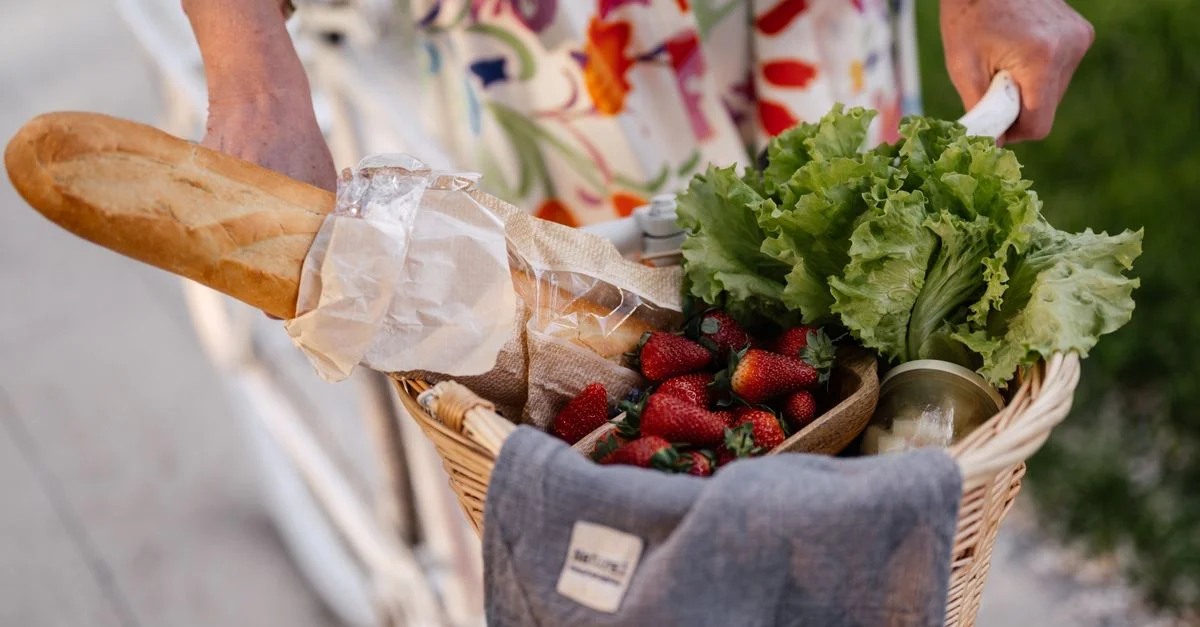 Une femme en robe à fleurs tient un panier rempli de pain, de fraises et de laitue sur son vélo.